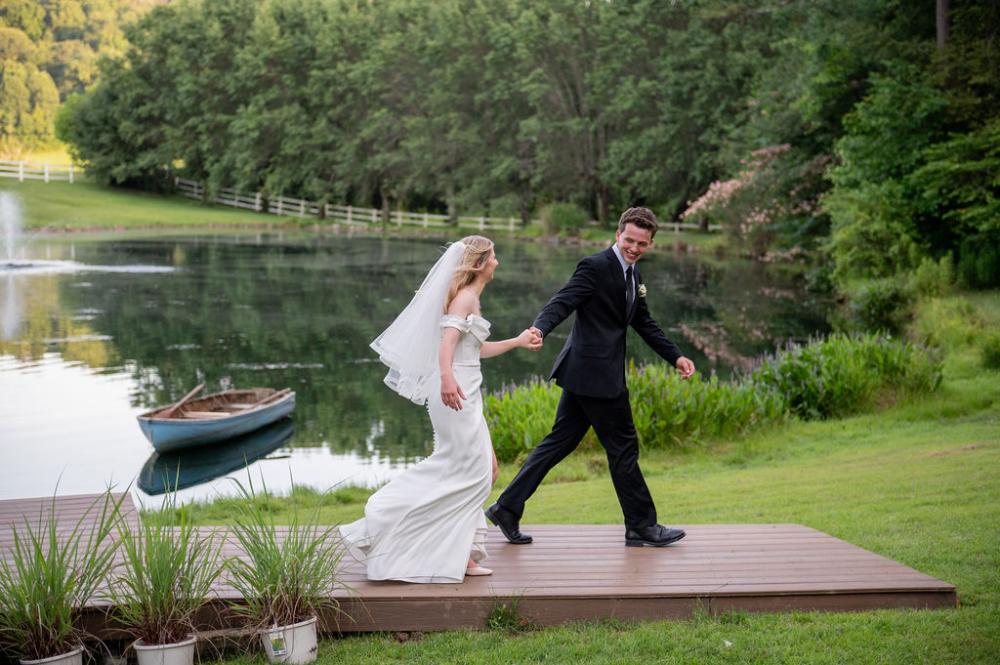 Bride and groom holding hands in front of a pond with a boat in it.